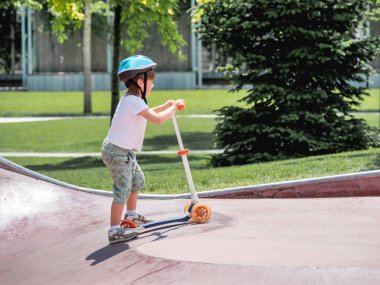 Cesur çocuk kaykay parkında scooter sürüyor. Şehir parkındaki özel beton kase yapıları. Yazın paten antrenmanı..