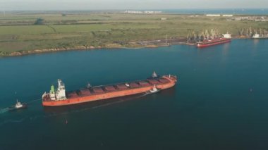 Aerial view of a large cargo ship leaving the harbor with tugboats at sunset.