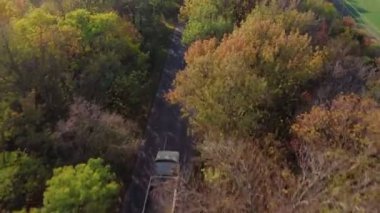Kırsal bir yol boyunca ilerleyen yalnız beyaz bir kamyonun havadan görünüşü. Bir sonbahar gününün ortasında yaprakları sararmış ağaçlar arasında..