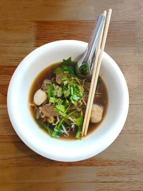 Tayland 'da bir restoranda tahta bir masaya yerleştirilmiş yemek çubukları ve kaşıklarla beyaz bir seramik kasede etli şehriye fotoğrafı..