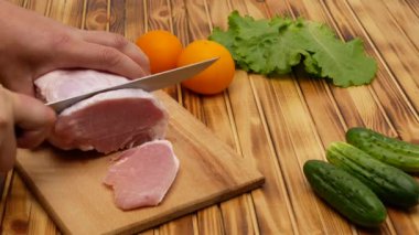 Cutting a piece of fresh meat, cutting meat in the kitchen. Next to the meat are cucumbers, lettuce and yellow tomatoes. Wooden background.