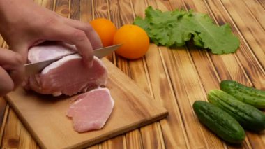 Cutting a piece of fresh meat, cutting meat in the kitchen. Next to the meat are cucumbers, lettuce and yellow tomatoes. Wooden background.