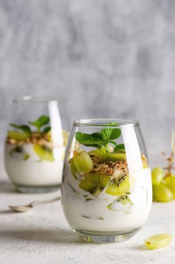 İki bardak sade yoğurt taze kivi meyveleri, beyaz üzümler, granola ve gri arkaplanlı nane yaprakları.. 
