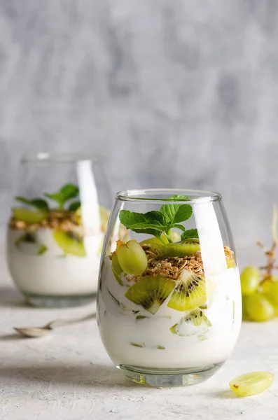 Two glasses of plain yoghurt with pieces of fresh kiwi fruits, white grapes, granola, and mint leaves with a grey background.