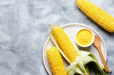 Üç mısır koçanı, bir kase mısır unu ve gri arka planda bir tabakta ahşap kaşık.. 