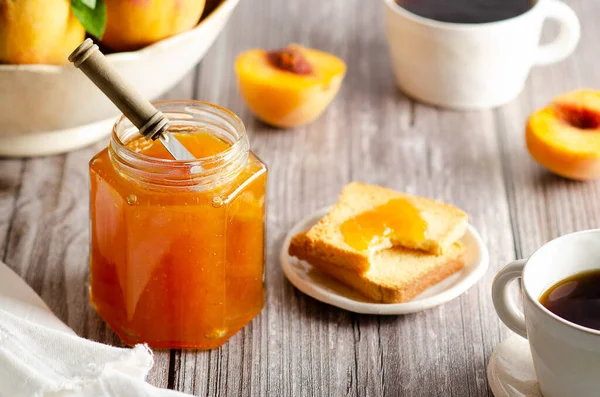 Kavanozda ev yapımı şeftali reçeli, biraz taze şeftali, iki fincan kahve ve birkaç tost, ahşap masa arkasında..