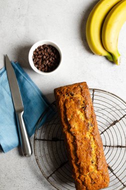 Çikolata parçacıklı muz ekmeği, iki muz, bir kasede çikolata parçacıkları ve gri arka planda mavi peçete.. 