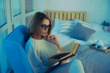 Boş zaman. Kız kitap okuyor, pencereye karşı koltukta oturuyor.