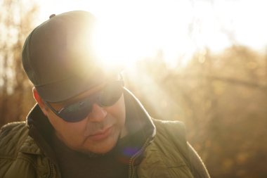 Şık kolsuz gömlekli yakışıklı bir hippi portresi, şık güneş gözlükleri ve güneşli park arka planında bir şapka. Yakışıklı sakallı adam aşağıya bakıyor..