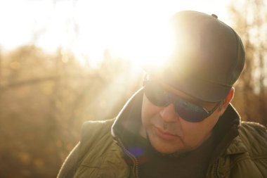 Şık kolsuz gömlekli yakışıklı bir hippi portresi, şık güneş gözlükleri ve güneşli park arka planında bir şapka. Yakışıklı sakallı adam aşağıya bakıyor..