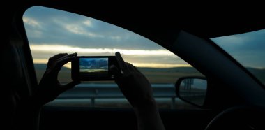 Erkek gezginin silueti arabanın içinde oturuyor, akıllı bir telefonla bakıyor ve fotoğraf çekiyor. Dağlarda güzel bir gün batımı. Fotoğraf çek.. 