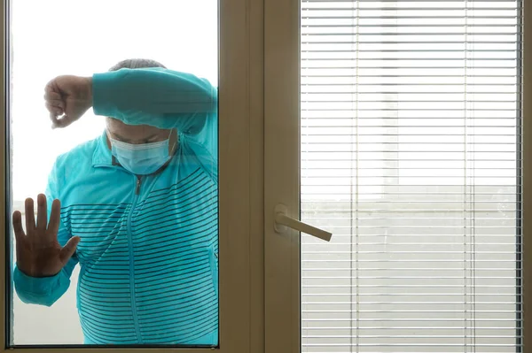 隔離された医療マスクの感染した男は 家の窓から通りを見ている 医療マスクの男は家にいる 家の中で自己分離 社会から隔離された隔離された — ストック写真