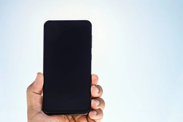 male hand holding a mobile cell phone isolated on blue sky background. black blank screen - mock up.