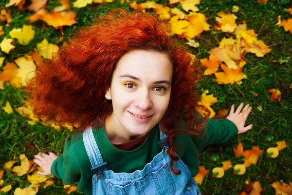 快乐的红头发少女坐在草地上 抬头看着摄像机 秋季公园 — 图库照片