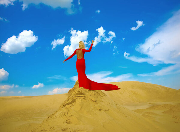 blonde woman posing in long red dress and  stand on sand desert against blue cloudy sky.  adult girl enjoying warm wind. Idea of of dry skin, Lack of water in body, outgoing time and youth. Back view