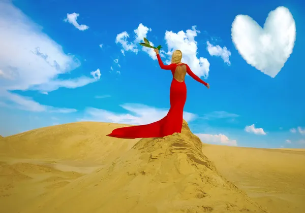 長い赤いドレスを着て砂漠の上に立つブロンドの女性 暖かい風を楽しんでいる大人の女の子 発信の時間と若者のアイデア バックビュー 青空の下で飛行機を打ち上げ — ストック写真