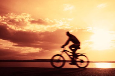 Bulanık hareket. Asfalt yolda bisiklete binen adam, yan görüş, arka planda deniz. Güneşli gün batımı bulutlu gökyüzü. güneş ışınları.