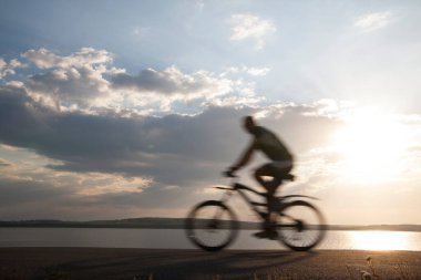 Bulanık hareket. Asfalt yolda bisiklete binen adam, yan görüş, arka planda deniz. Güneşli gün batımı bulutlu gökyüzü. güneş ışınları.