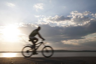 Bulanık hareket. Asfalt yolda bisiklete binen adam, yan görüş, arka planda deniz. Güneşli gün batımı bulutlu gökyüzü. güneş ışınları.
