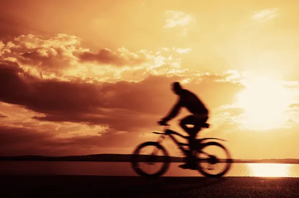 模糊的运动 人骑自行车在柏油路上 背景中的大海 夕阳西下多云的天空 太阳光 — 图库照片