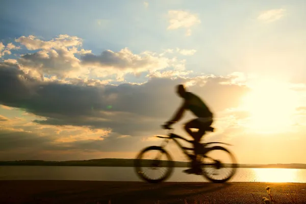 ぼやけた動きだ アスファルトの道路 サイドビュー バックグラウンドで海での男の自転車 晴れた日没曇天 太陽光線 — ストック写真