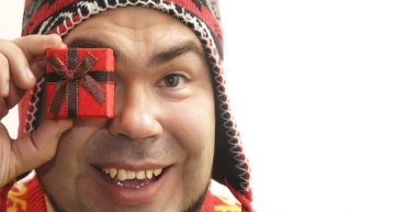 Arkasında hediye kutusu tutan, büyük mutlu gözlerle kameraya bakan kırmızı kazak giyen ve kırmızı örgü şapkalı bir adam. Mutlu yıllar ve mutlu Noeller..