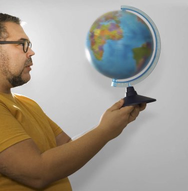 Genç, beyaz sakallı, coğrafya öğrencisi, sarı tişörtlü, öfkeli adam Dünya 'nın dünyasını ele geçirdi. Erkek, gri arkaplanda izole edilmiş dünya haritasına bakıyor. Stüdyo çekimi. 