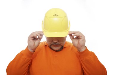 Tişörtlü adam sarı kaskın güneşliğinin altından bakıyor..