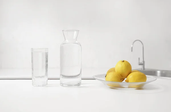 新鲜的柠檬和玻璃瓶 清澈的水屹立在现代室内装饰的木制桌子上 与厨房的白色碗橱和台面形成模糊的背景 纯净水水A — 图库照片