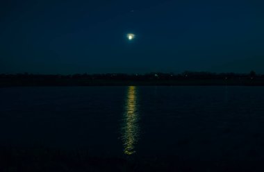 Beautiful landscape view of a glowing moon beginning its rise over Lake. A pale yellow aura surrounds the moon and a strip of light reflects on the water.