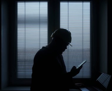 silhouette of architect or foreman in helmet working with laptop and mobile phone on the apartment drawings on blue window with blinds background.
