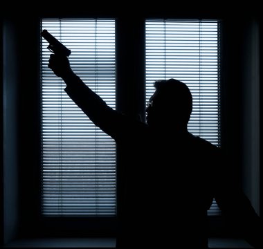 A man with a gun is standing by the window blind, ready to protect himself from another bad people, crime scene conceptual. silhouette of an armed man behind blinds