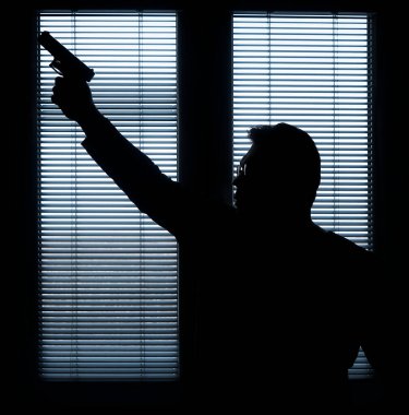 A man with a gun is standing by the window blind, ready to protect himself from another bad people, crime scene conceptual. silhouette of an armed man behind blinds