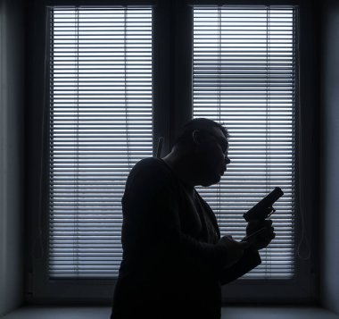 A man with a gun is standing by the window blind, ready to protect himself from another bad people, crime scene conceptual. silhouette of an armed man behind blinds.