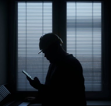 silhouette of architect or foreman in helmet working with laptop and mobile phone on the apartment drawings on blue window with blinds background.