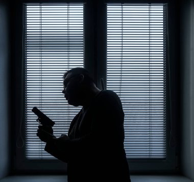 A man with a gun is standing by the window blind, ready to protect himself from another bad people, crime scene conceptual. silhouette of an armed man behind blinds.