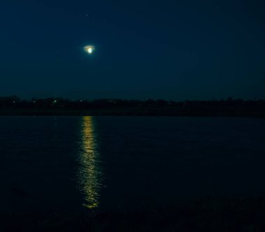 Beautiful landscape view of a glowing moon beginning its rise over Lake. A pale yellow aura surrounds the moon and a strip of light reflects on the water.