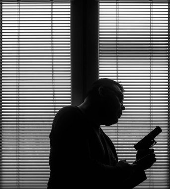 A man with a gun is standing by the window blind, ready to protect himself from another bad people, crime scene conceptual. silhouette of an armed man behind blinds.
