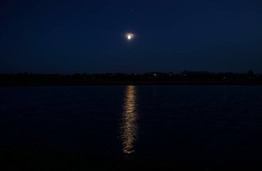 Beautiful landscape view of a glowing moon beginning its rise over Lake. A pale yellow aura surrounds the moon and a strip of light reflects on the water.