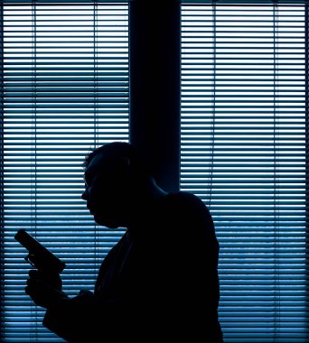 A man with a gun is standing by the window blind, ready to protect himself from another bad people, crime scene conceptual. silhouette of an armed man behind blinds.