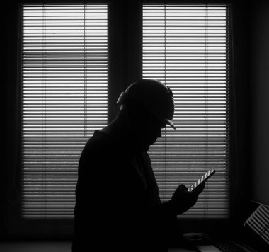 silhouette of architect or foreman in helmet working with laptop and mobile phone on the apartment drawings on blue window with blinds background.