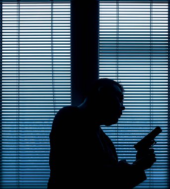 A man with a gun is standing by the window blind, ready to protect himself from another bad people, crime scene conceptual. silhouette of an armed man behind blinds.