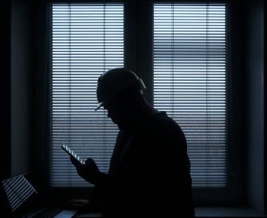 silhouette of architect or foreman in helmet working with laptop and mobile phone on the apartment drawings on blue window with blinds background.