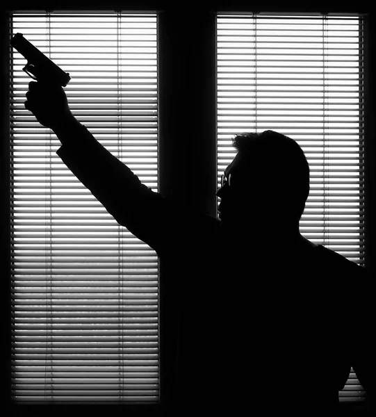 A man with a gun is standing by the window blind, ready to protect himself from another bad people, crime scene conceptual. silhouette of an armed man behind blinds