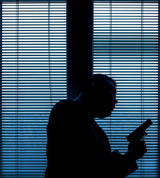 A man with a gun is standing by the window blind, ready to protect himself from another bad people, crime scene conceptual. silhouette of an armed man behind blinds.