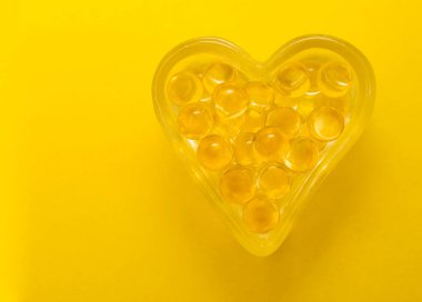 Sarı arka planda izole edilmiş kalp şeklinde morina yağı Omega 3 jel kapsülleri. Kalp şeklinde gözlük kutusu..