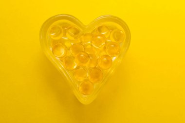 Sarı arka planda izole edilmiş kalp şeklinde morina yağı Omega 3 jel kapsülleri. Kalp şeklinde gözlük kutusu..