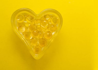 Sarı arka planda izole edilmiş kalp şeklinde morina yağı Omega 3 jel kapsülleri. Kalp şeklinde gözlük kutusu..