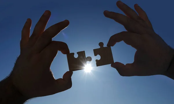 two hands connecting couple puzzle pieces against sky background. Jigsaw wooden puzzle against sun rays. symbol of association and connection. business strategy