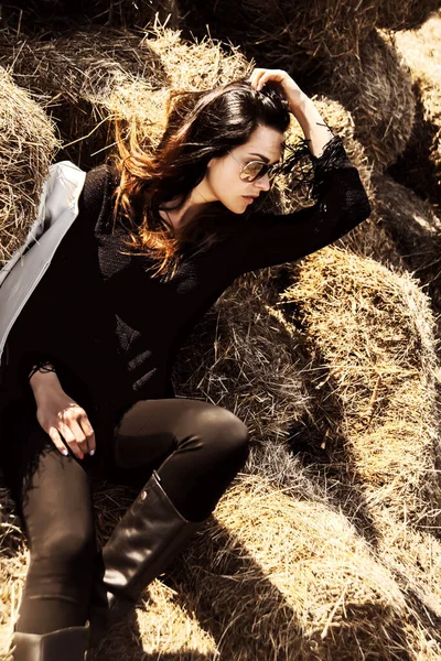 黒のスタイリッシュな服と革のハイブーツで美しいブルネットの女の子の肖像画 女性は村の農場の干し草の山の上に座っている 暖かい日差しを楽しむ女 — ストック写真